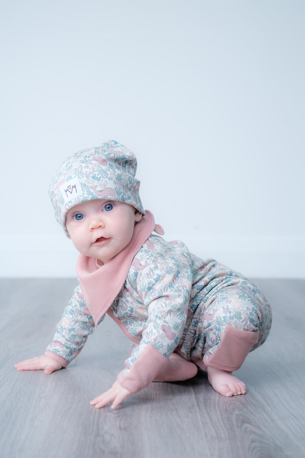 A baby crawling on a grey floor against a light grey background, wearing a floral print one-piece outfit with dusty pink accents and a matching beanie hat.