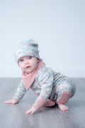 A baby crawling on a grey floor against a light grey background, wearing a floral print one-piece outfit with dusty pink accents and a matching beanie hat.
