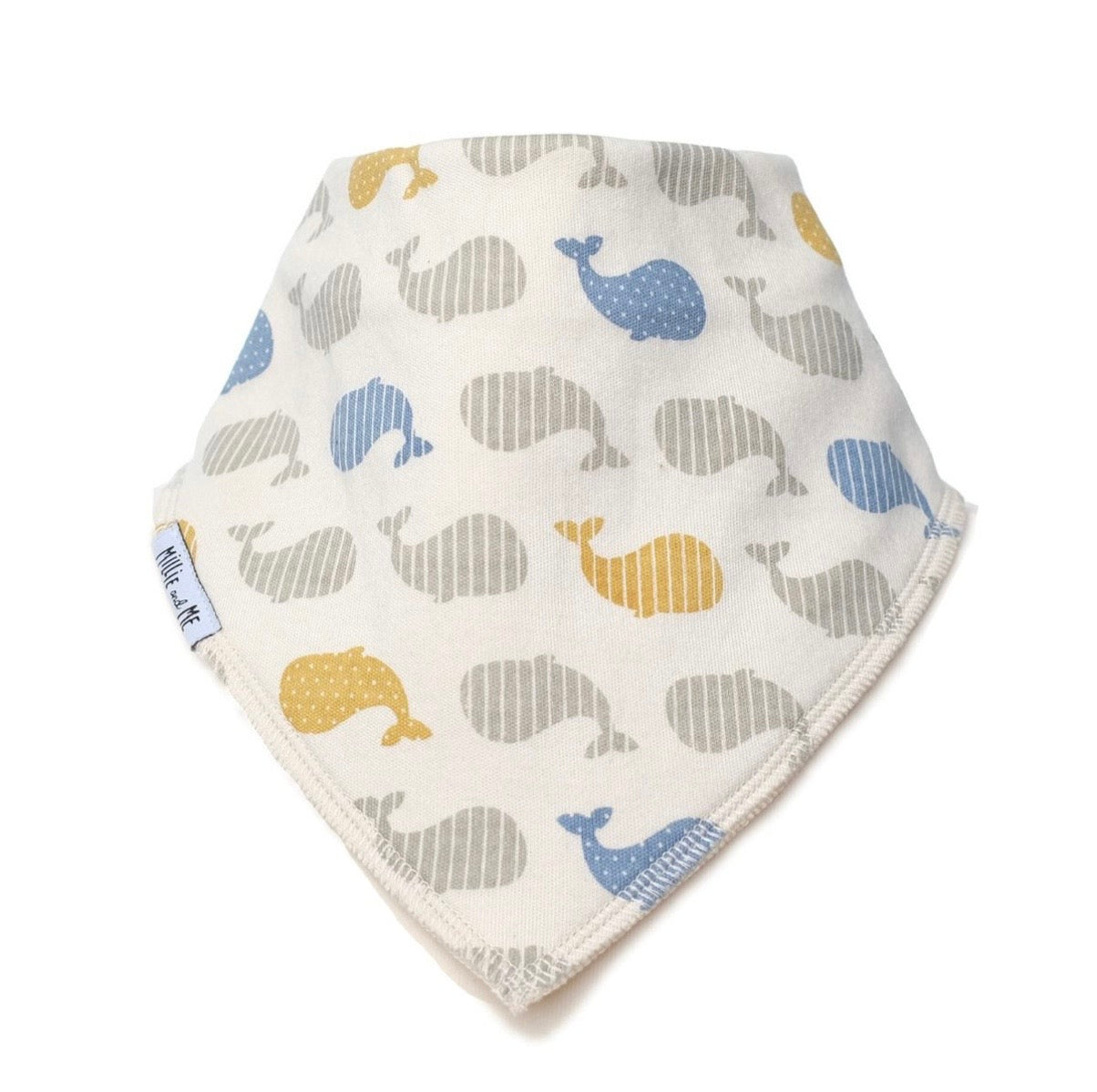 A triangular baby bandana bib with an off-white background, featuring a pattern of stylised whales in blue, grey, and mustard yellow. A small white fabric tag on the left edge displays the brand name 'Millie and Me'.