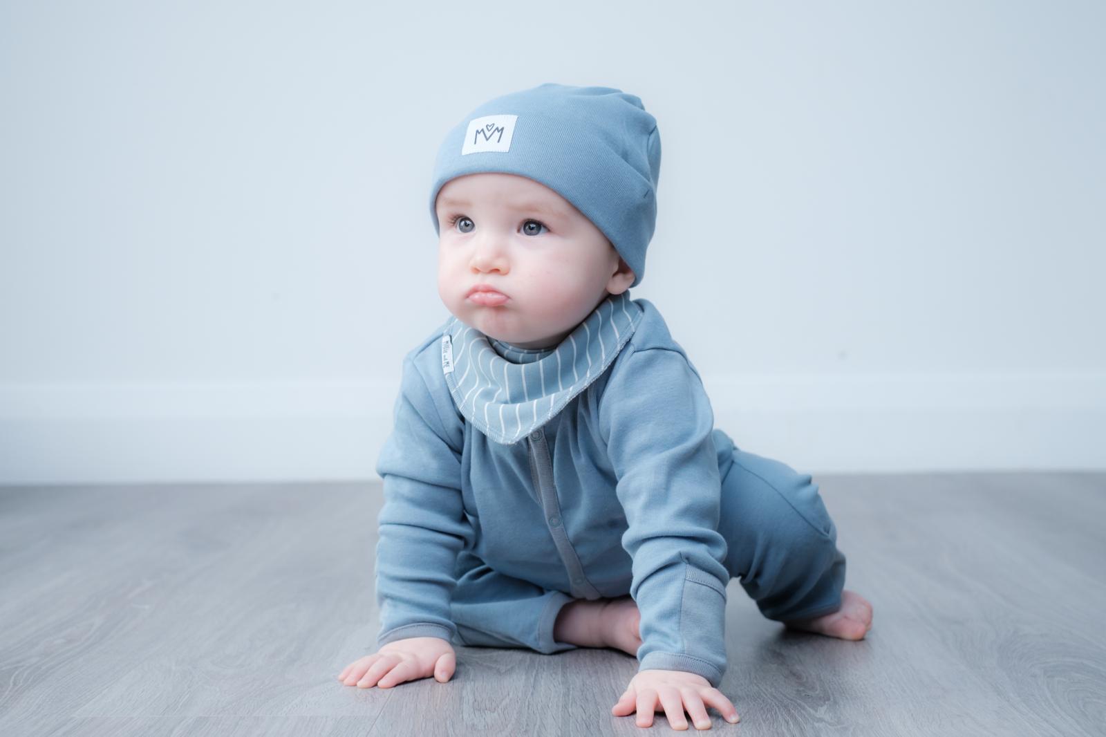 A baby crawling on a gray wood floor against a light grey wall, wearing a dusty blue long-sleeve romper, matching beanie hat, and a striped bib.