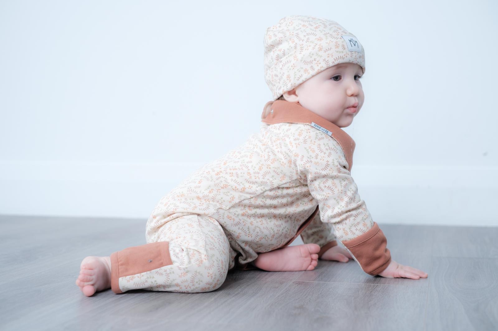 A baby wearing a beige and white patterned long-sleeve romper with brown cuffs and a matching beanie hat, crawling on a grey floor against a white wall.