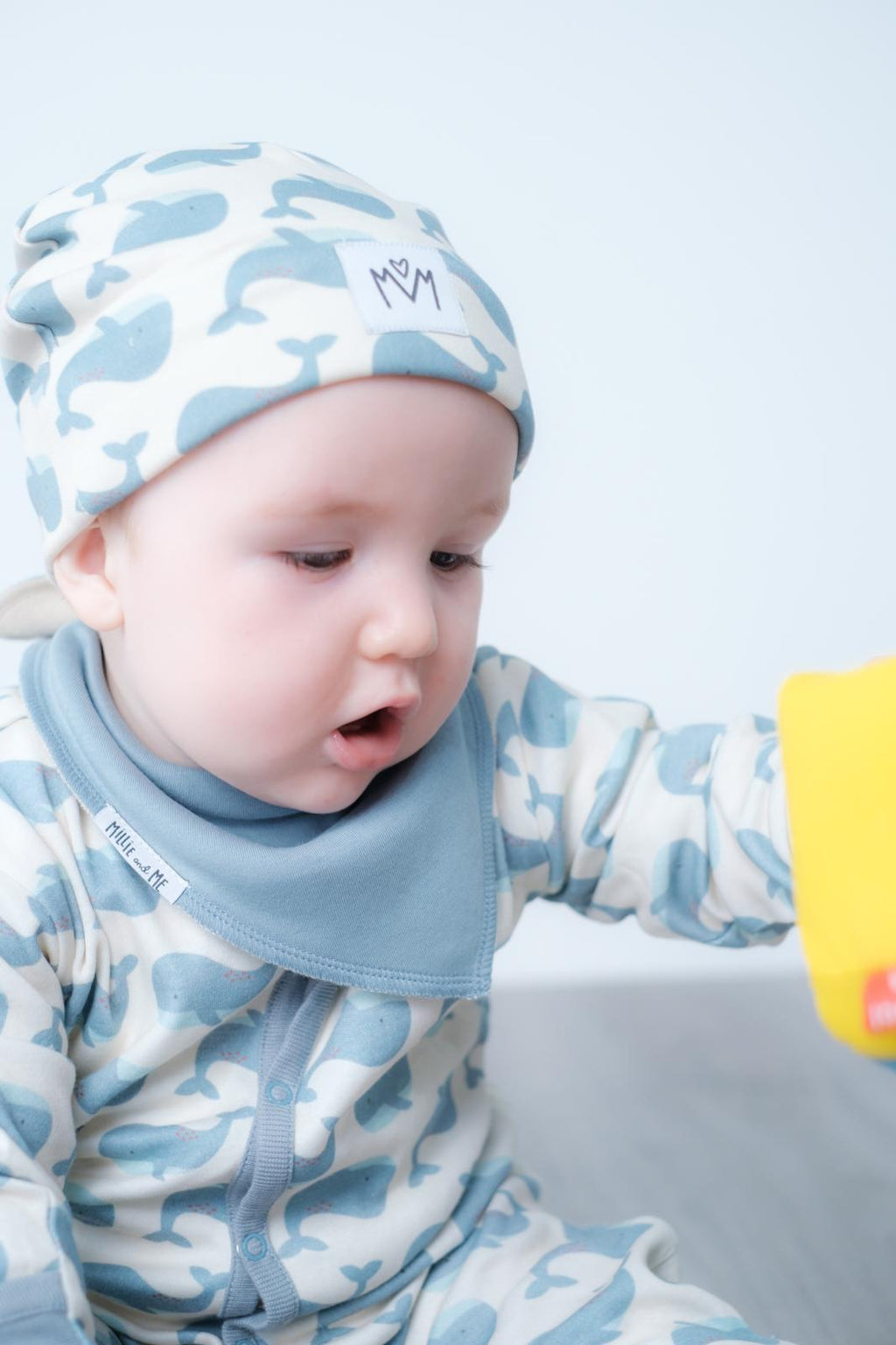 A baby wearing a light blue and white camouflage patterned footed sleeper with a matching beanie and a light blue bib. The baby is looking down and to the side, with a small yellow toy visible on the right edge.
