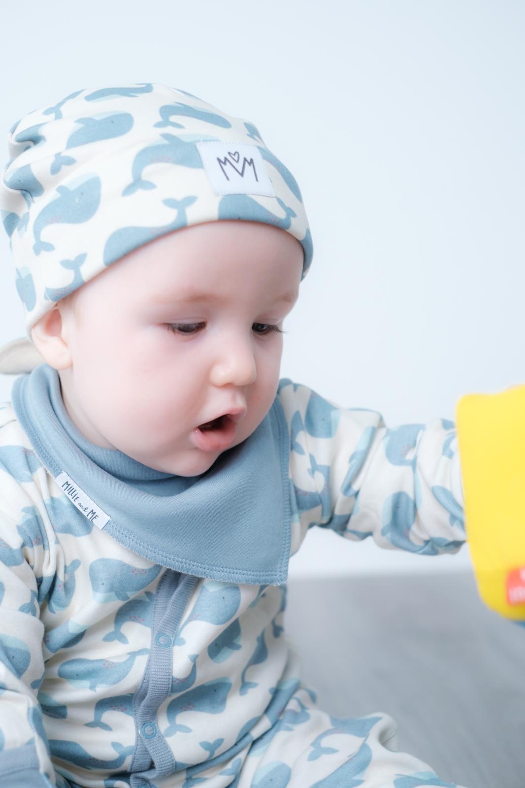 A baby wearing a light blue and white camouflage patterned footed sleeper with a matching beanie and a light blue bib. The baby is looking down and to the side, with a small yellow toy visible on the right edge.