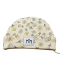 A cream-colored baby beanie hat featuring a daisy pattern with white petals, yellow centers, and gray outlines. It has a white fabric patch on the front with a black hand-drawn logo.