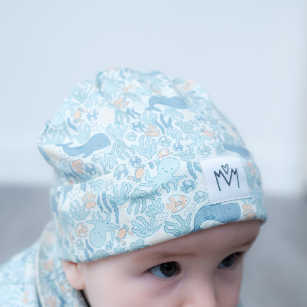 A close-up of a baby wearing a light blue patterned beanie with sea creatures and a small white patch with the letters 'M M'.