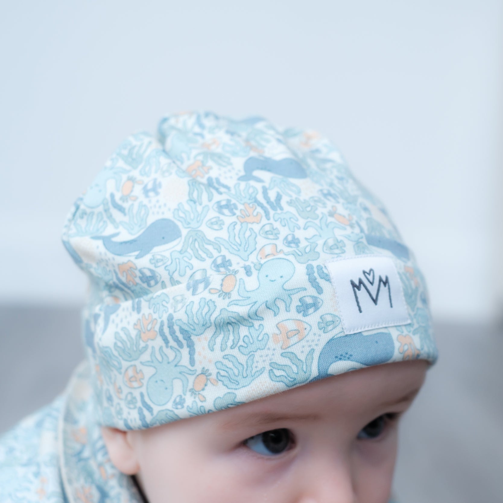 A close-up of a baby wearing a light blue patterned beanie with sea creatures and a small white patch with the letters 'M M'.
