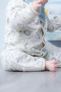 A baby sitting on a grey floor wearing a cream-coloured footless sleeper with a black line art animal pattern, paired with a light blue striped bib.