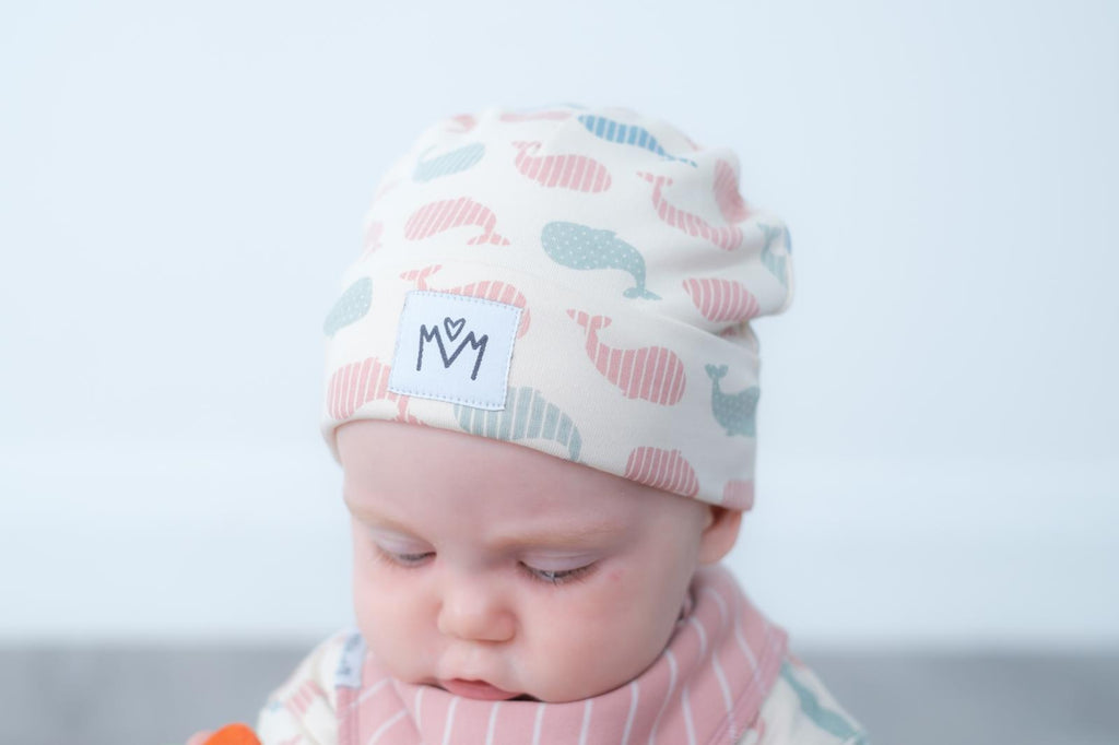 A baby wearing a soft knit beanie hat with a whale pattern in pink, blue, and grey on a cream background, along with a matching striped bib. The hat features a white patch with the brand logo.