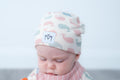 A baby wearing a soft knit beanie hat with a whale pattern in pink, blue, and grey on a cream background, along with a matching striped bib. The hat features a white patch with the brand logo.