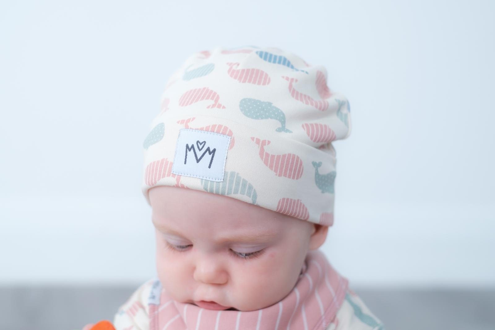 A baby wearing a soft knit beanie hat with a whale pattern in pink, blue, and grey on a cream background, along with a matching striped bib. The hat features a white patch with the brand logo.