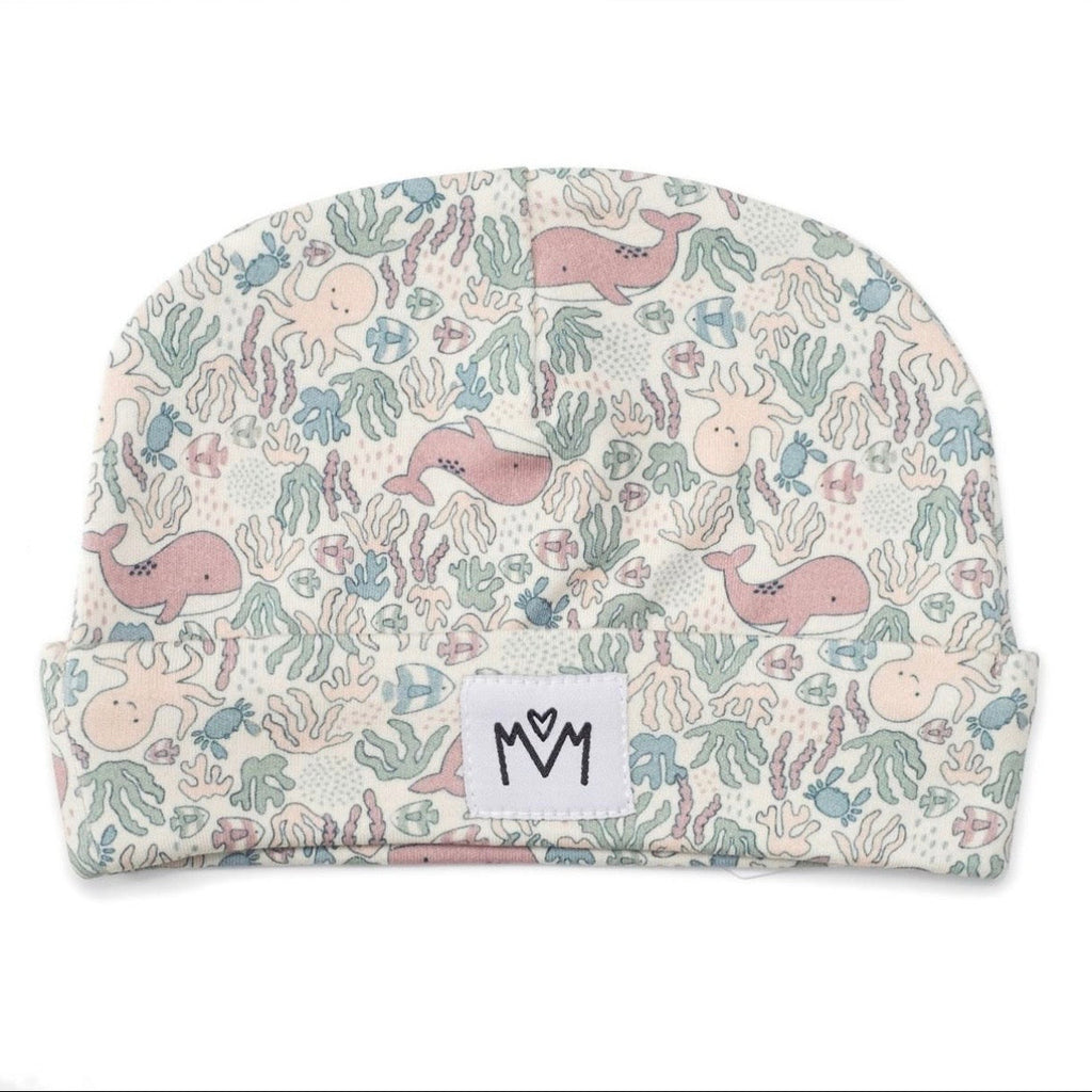 A baby beanie hat featuring a pastel underwater pattern with whales, octopus, and sea plants on a cream background, with a white fabric label with logo on the front cuff.