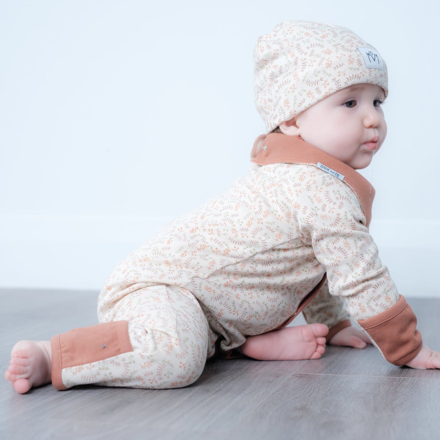 A baby wearing a beige and white patterned long-sleeve romper with brown cuffs and a matching beanie hat, crawling on a gray floor against a white wall.