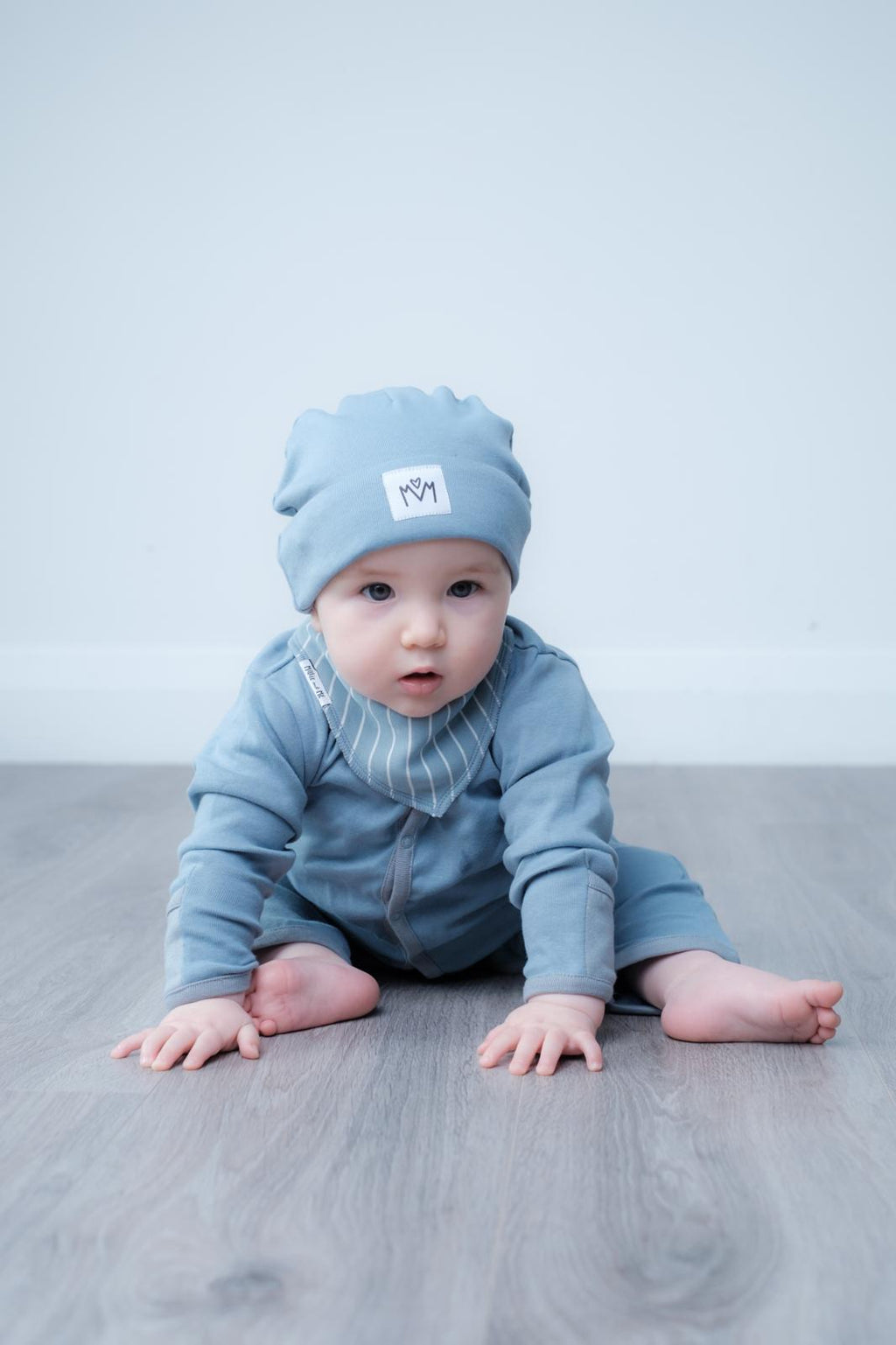 A baby crawling on a gray wooden floor against a light gray wall, wearing a light blue knit beanie with a small white square patch and a matching light blue long-sleeve outfit with a striped bib.