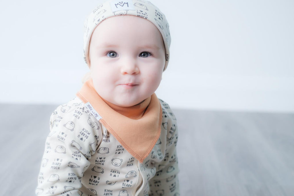 A baby sitting on a grey wood floor against a light grey background, wearing a cream-coloured long-sleeve onesie and matching bonnet with a black print, and a peach-coloured bib.