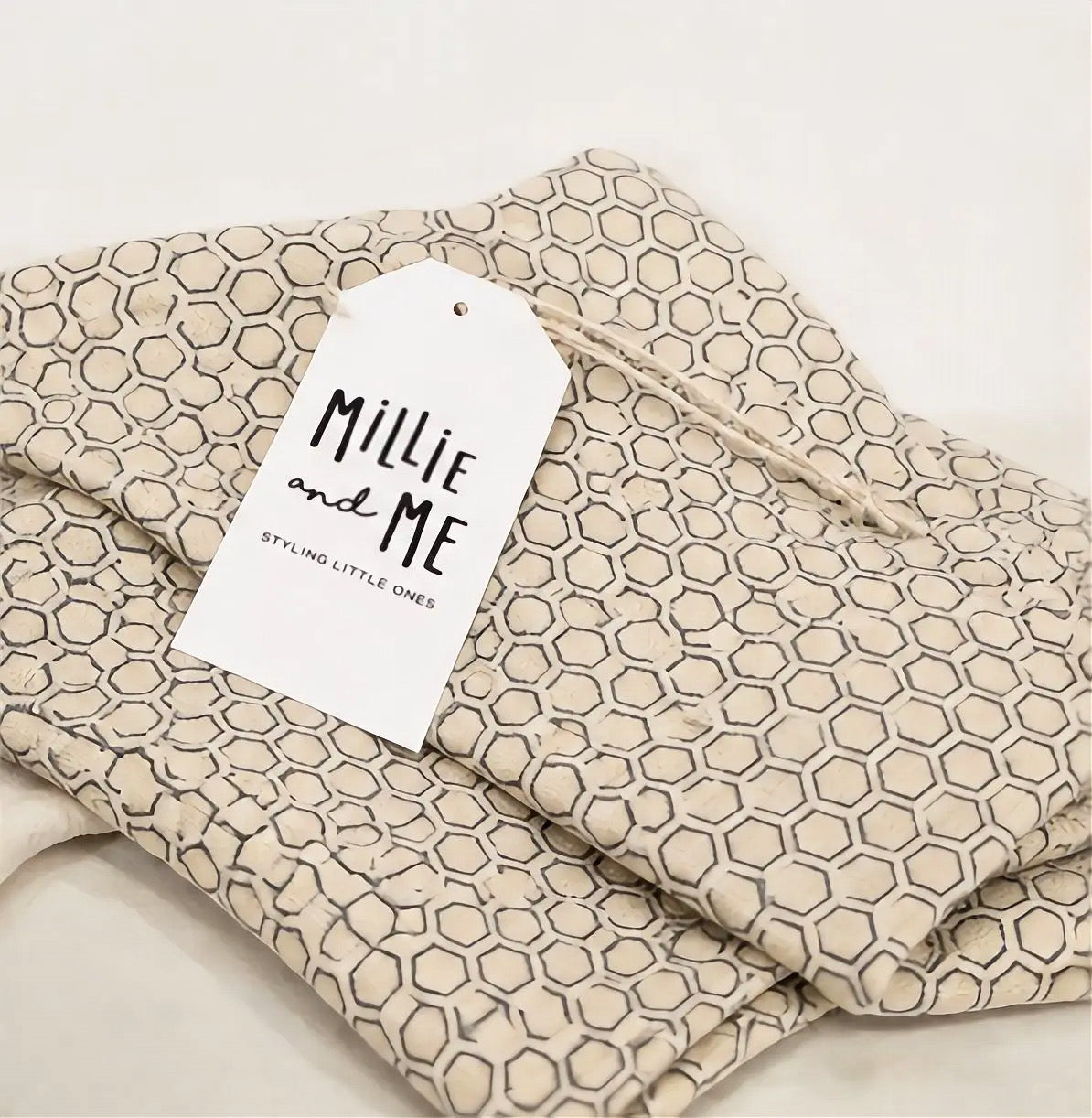 Folded beige fabric garment with a repeating blue geometric hexagon pattern, shown with a white paper tag reading Millie and Me.