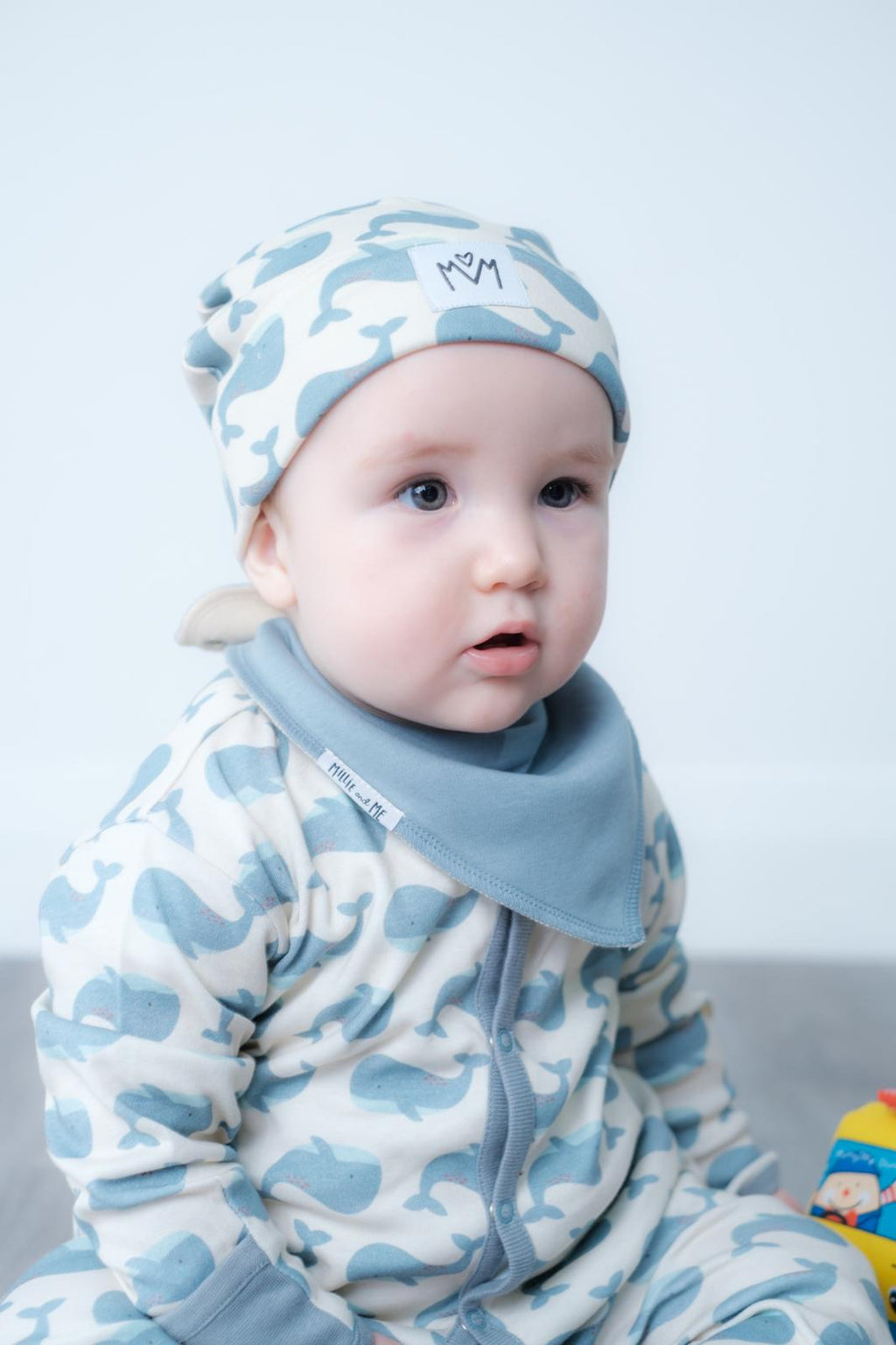 A baby wearing a light blue and white camouflage patterned one-piece outfit with a matching beanie hat and a solid light blue bib, sitting against a plain light grey background.