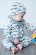 A baby sitting on a grey wooden floor wearing a light blue and white whale-patterned zip-up onesie and matching beanie hat, holding a yellow toy bus.