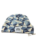 A folded fabric beanie hat with a geometric triangle pattern in shades of navy, slate blue, and cream. A white square patch on the front features the stylised logo with a small heart between the letters.