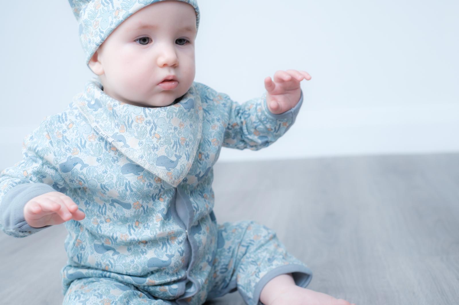 A baby wearing a light blue patterned one-piece jumpsuit with a matching bonnet and bib, sitting on a grey floor against a white background.