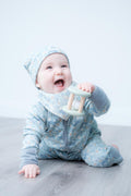 A smiling baby wearing a light blue floral-patterned footed onesie and matching bonnet sits on a grey wood floor, holding a pale green wooden teething toy.