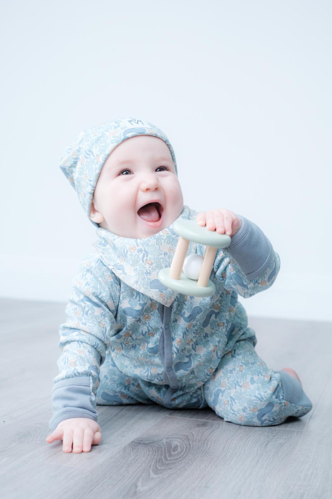 A smiling baby wearing a light blue floral-patterned footed onesie and matching bonnet sits on a grey wood floor, holding a pale green wooden teething toy.