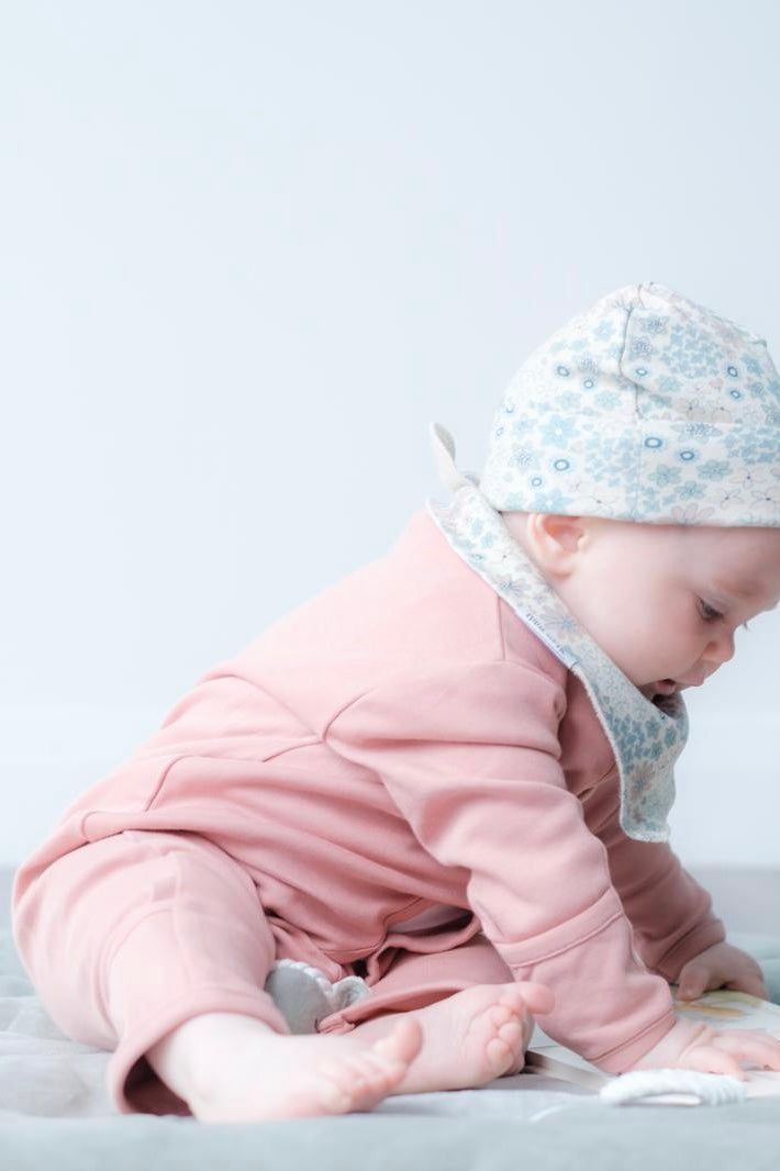 A baby wearing a dusty pink one-piece outfit and a light blue and white floral bonnet, crawling on a light gray blanket against a white background.