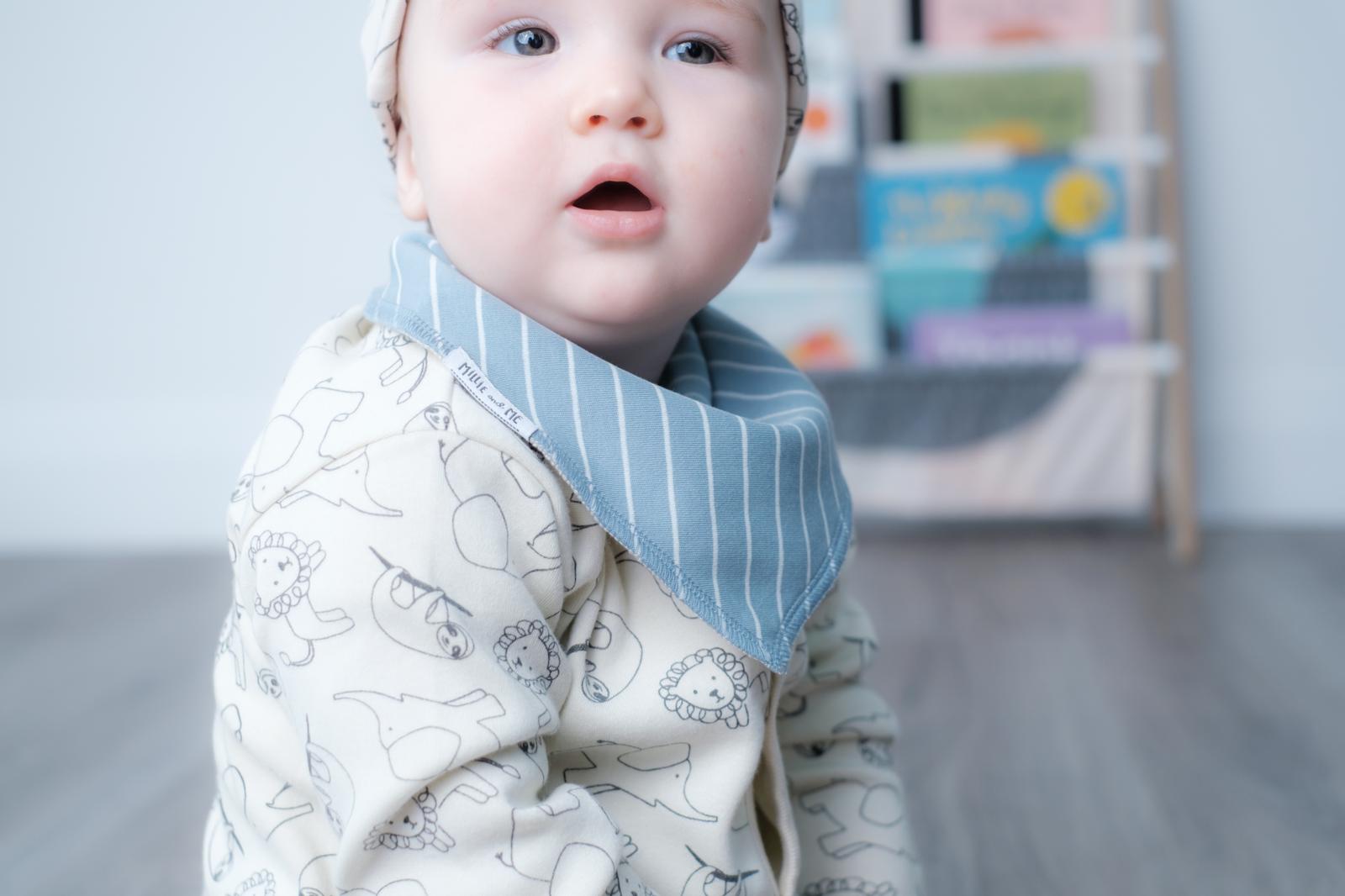 A baby wearing a cream-colored long-sleeve onesie with a subtle animal line pattern and a light blue striped bib, sitting on a wooden floor in a softly blurred nursery setting.