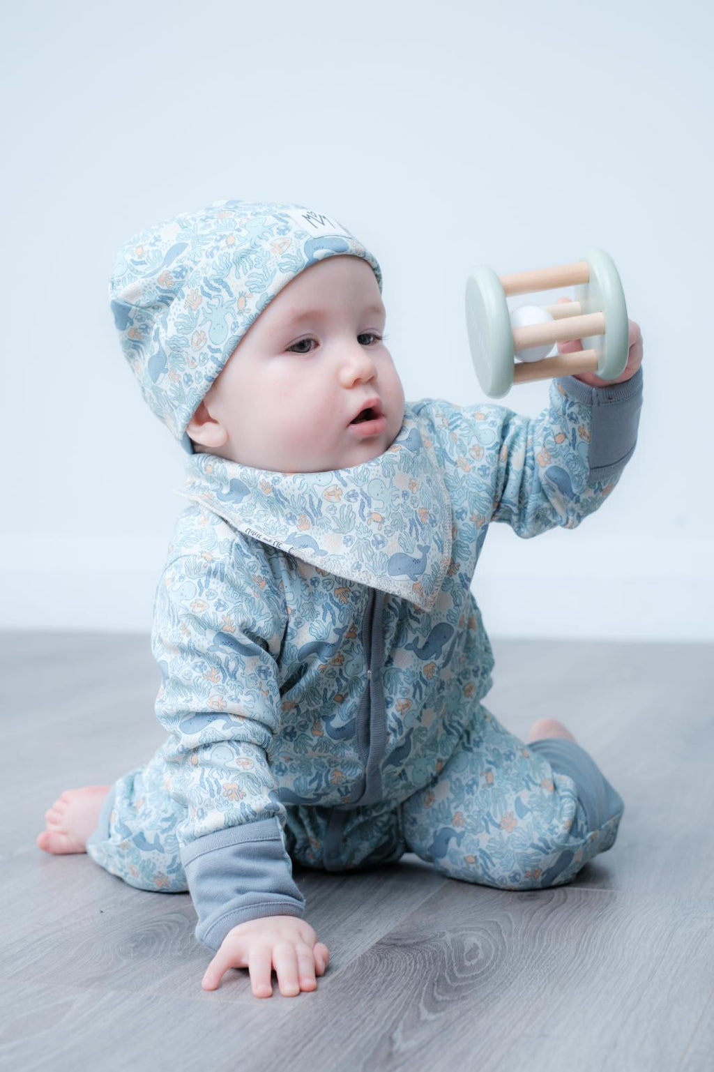 A baby wearing a light blue floral print one-piece outfit with a matching bonnet and bib, sitting on a grey floor and holding a wooden toy rattle.