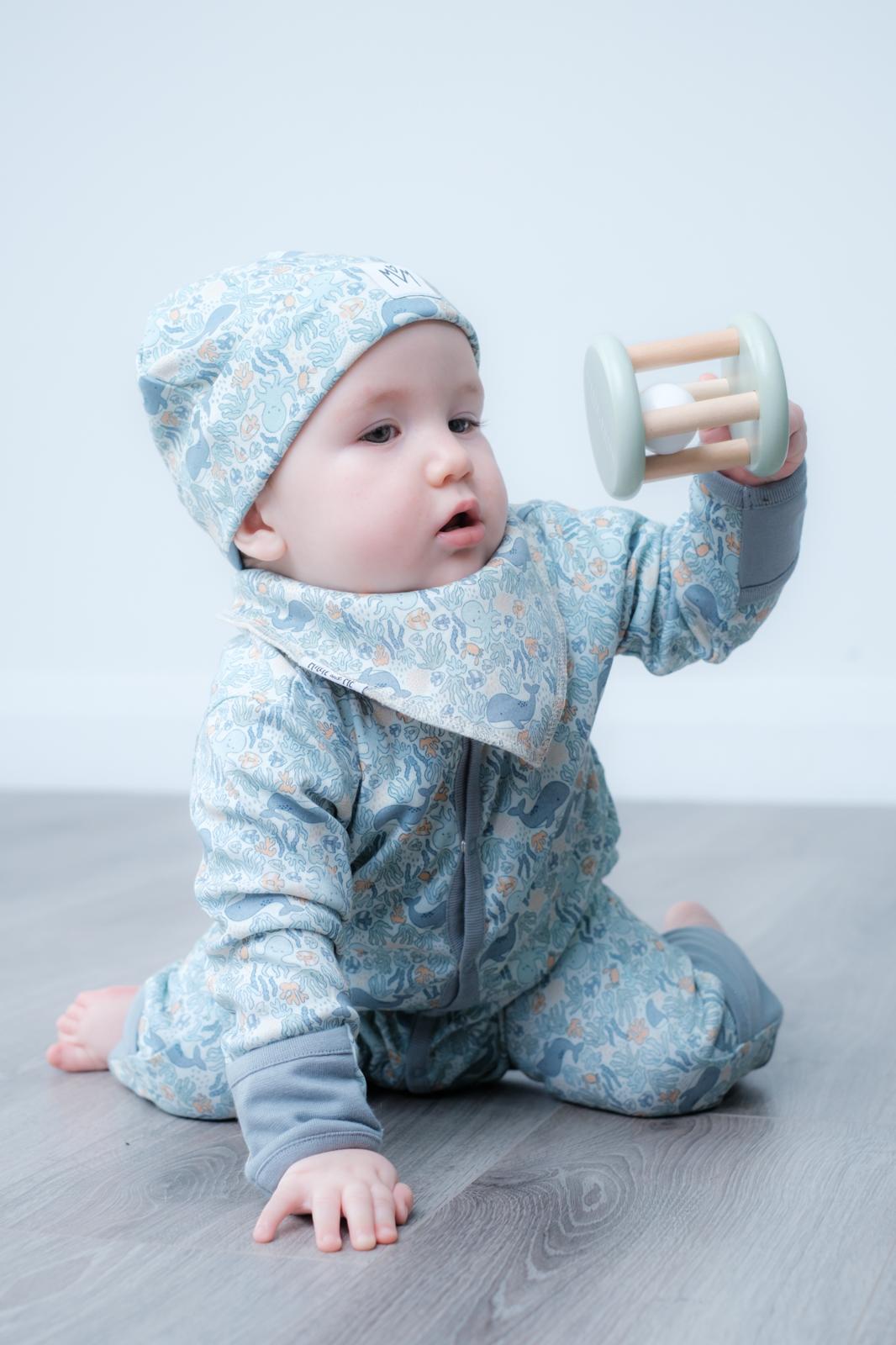 A baby wearing a light blue floral print one-piece outfit with a matching bonnet and bib, sitting on a grey floor and holding a wooden toy rattle.