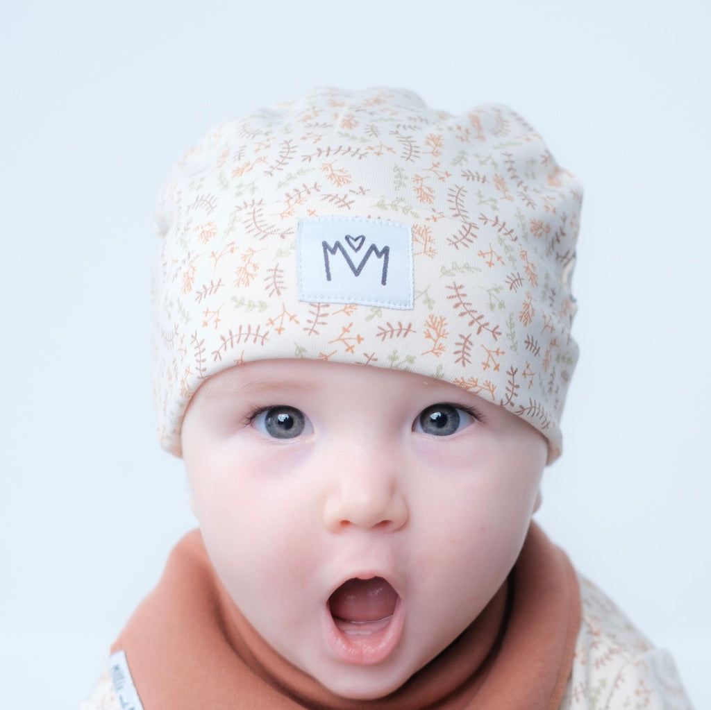 A close-up of a baby wearing a cream-colored knit beanie with a delicate tan botanical pattern and a white patch featuring the black embroidered letters 'M' and 'M' with a heart. The baby is also wearing a soft brown bib against a clean, light gray background.