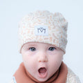 A close-up of a baby wearing a cream-colored knit beanie with a delicate tan botanical pattern and a white patch featuring the black embroidered letters 'M' and 'M' with a heart. The baby is also wearing a soft brown bib against a clean, light gray background.