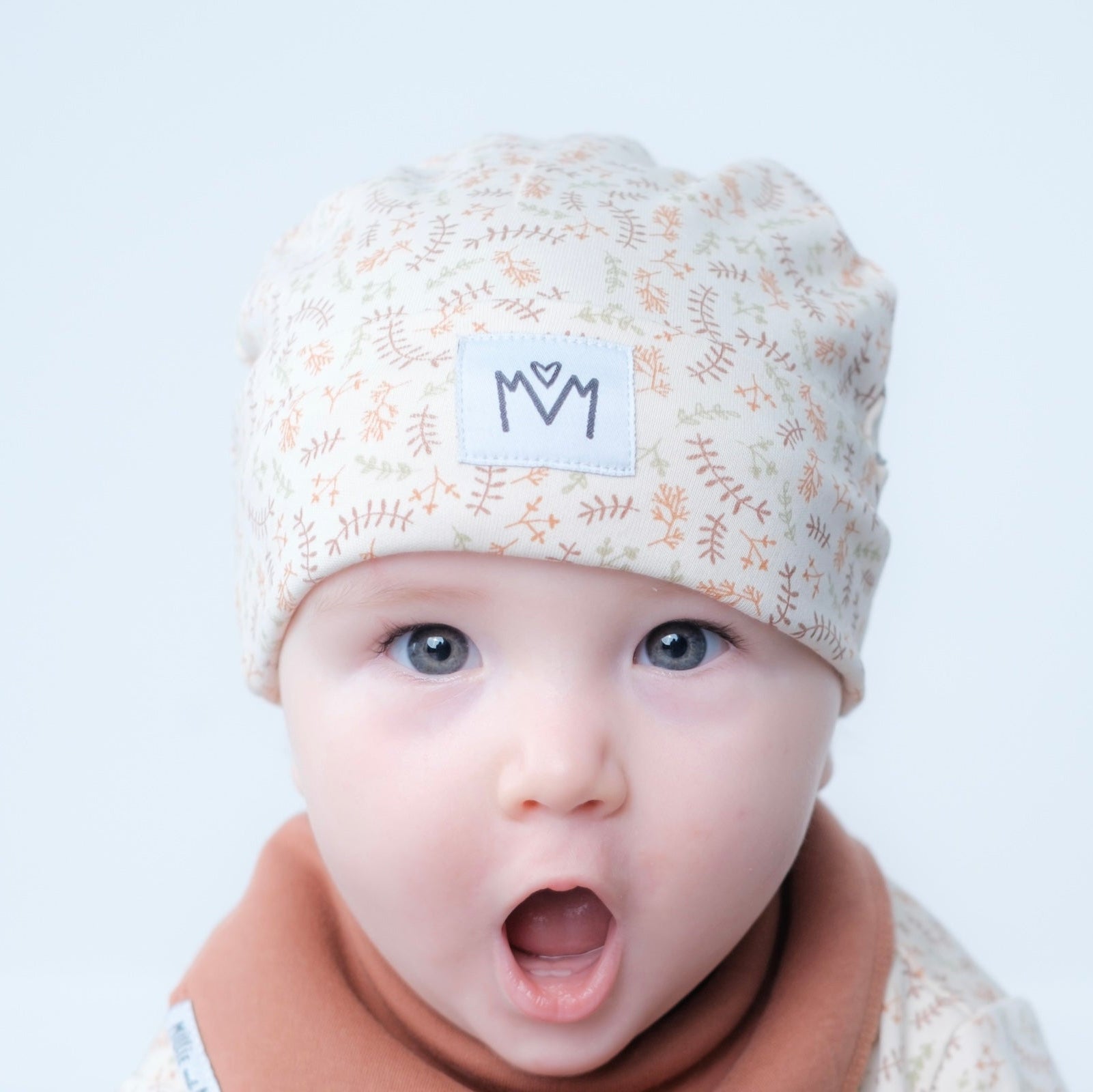 A close-up of a baby wearing a cream-colored knit beanie with a delicate tan botanical pattern and a white patch featuring the black embroidered letters 'M' and 'M' with a heart. The baby is also wearing a soft brown bib against a clean, light gray background.
