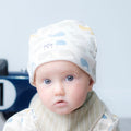 A baby wearing a cream-coloured whale-patterned beanie and a matching striped bib, looking directly at the camera with a blue toy race car blurred in the background.