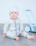 A baby sitting on a grey wooden floor wearing a light blue and white striped one-piece romper and matching beanie hat, with a light blue toy walker in the background.