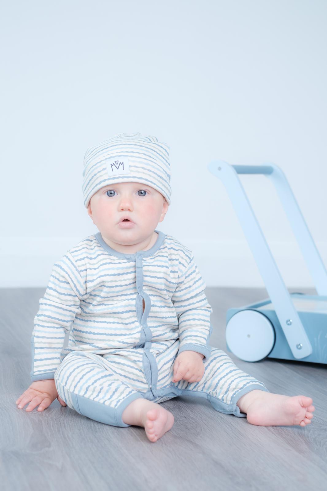 A baby sitting on a grey wooden floor wearing a light blue and white striped one-piece romper and matching beanie hat, with a light blue toy walker in the background.