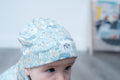 A close-up of a baby wearing a light blue patterned beanie with sea creatures and a small white patch with black letters, photographed indoors with a blurred background.