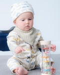 A baby wearing a cream-coloured striped and cloud-patterned onesie and matching hat sits on a grey floor, holding a clear cylindrical toy with colourful spiral rings.