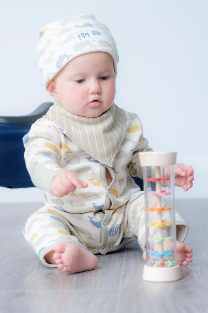 A baby wearing a cream-coloured striped and cloud-patterned onesie and matching hat sits on a grey floor, holding a clear cylindrical toy with colourful spiral rings.