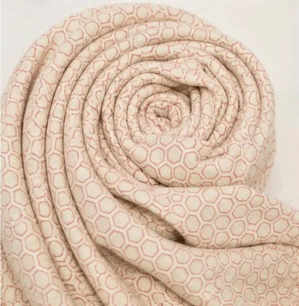 A soft, rolled fabric item, likely a scarf or throw blanket, featuring a repeating geometric hexagon pattern in dusty pink on a warm cream background.