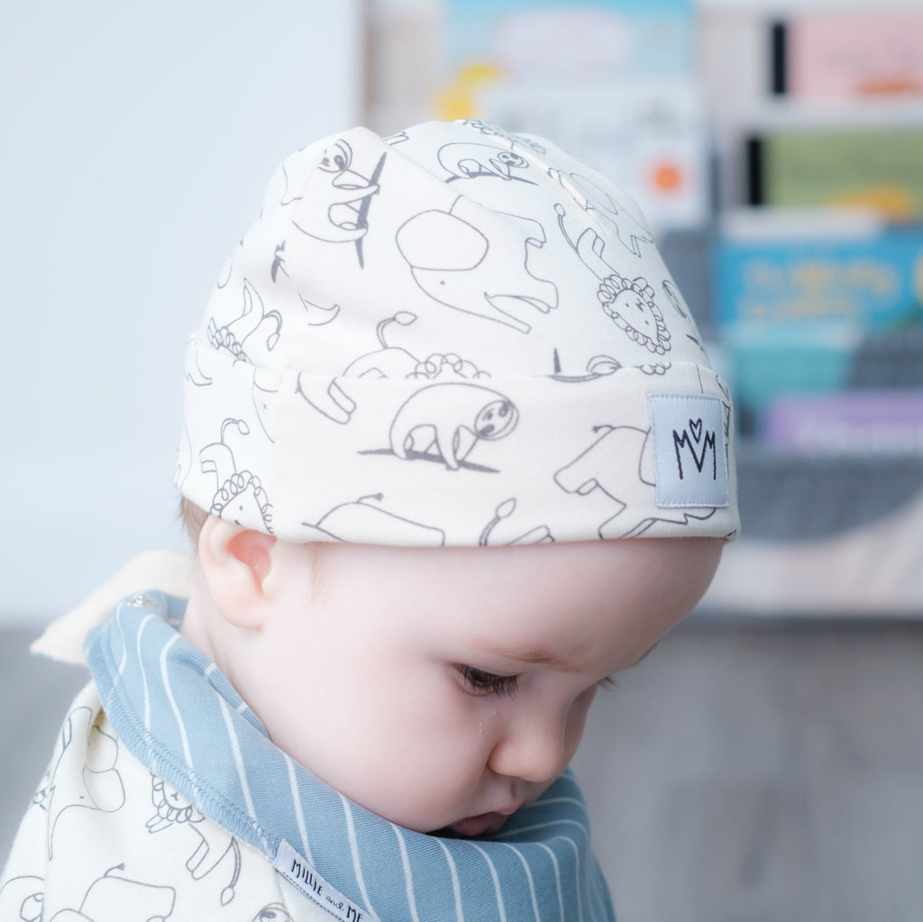 A baby wearing a white knit beanie with black line drawings of animals and a blue striped bib, captured in a softly lit nursery setting.