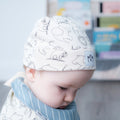 A baby wearing a white knit beanie with black line drawings of animals and a blue striped bib, captured in a softly lit nursery setting.