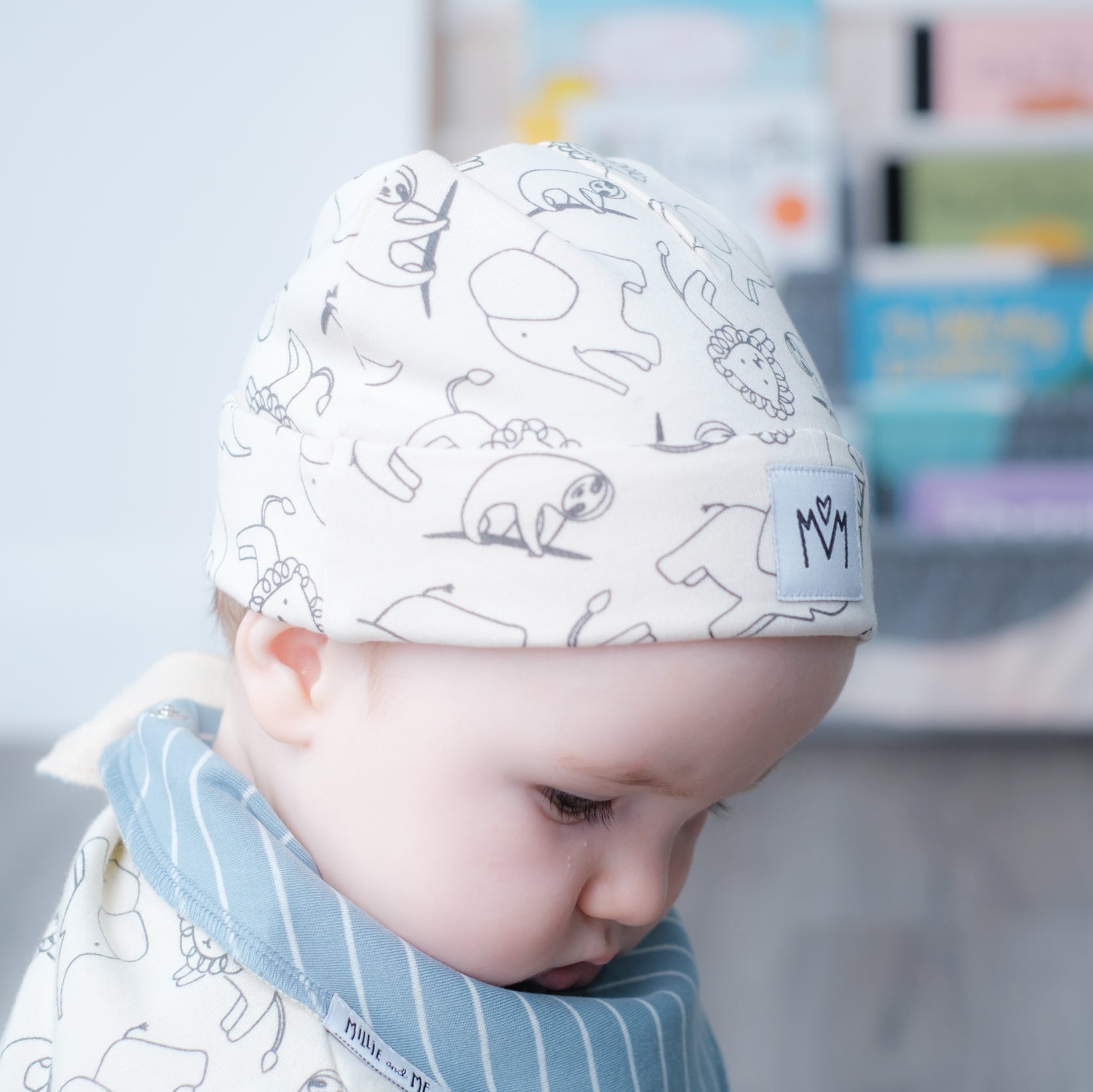 A baby wearing a white knit beanie with black line drawings of animals and a blue striped bib, captured in a softly lit nursery setting.