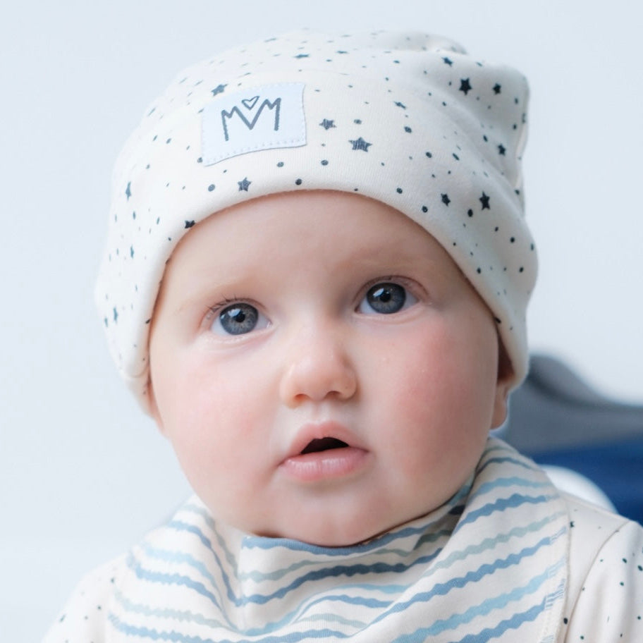 A close-up of a baby wearing a cream knit beanie with small dark star prints and a matching striped bib, against a soft light gray background.