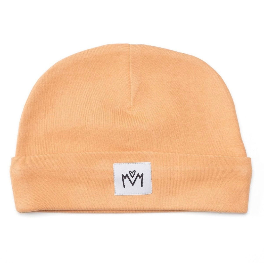 A peach-colored knit beanie hat with a folded cuff and a small white square patch featuring a black hand-drawn logo, shown on a plain white background.