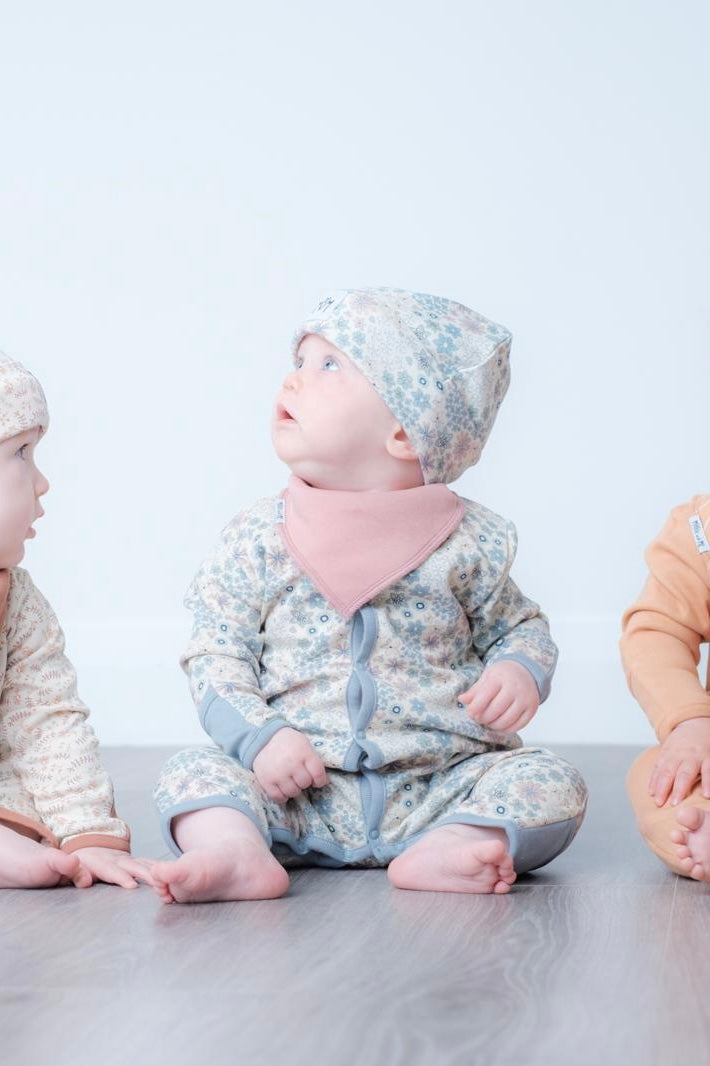A baby sitting on a grey floor wearing a light blue and white floral print footie, a pink bib, and a matching floral bonnet. Two other babies are partially visible on either side.