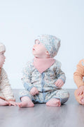 A baby sitting on a grey floor wearing a light blue and white floral print footie, a pink bib, and a matching floral bonnet. Two other babies are partially visible on either side.