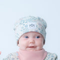 A baby wearing a light blue and white floral patterned beanie with a white patch that says 'MM' and a pink bib, photographed against a clean white background.