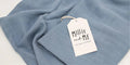 A light blue textured fabric item, likely a scarf or wrap, draped on a white surface with a white paper tag attached that reads 'Millie and Me'.