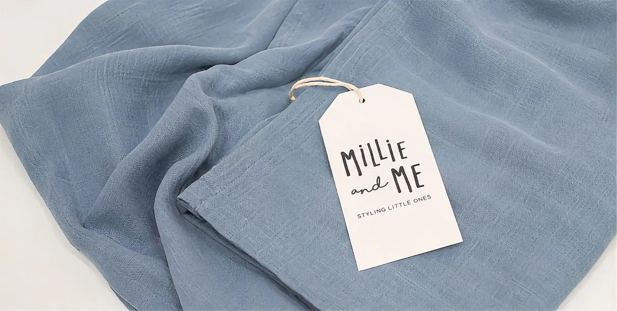 A light blue textured fabric item, likely a scarf or wrap, draped on a white surface with a white paper tag attached that reads 'Millie and Me'.