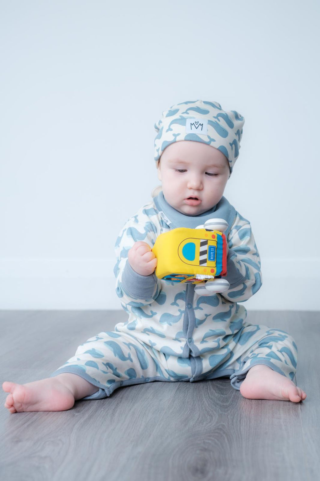 A baby sitting on a grey wood floor wearing a light blue and white camouflage print one-piece outfit and matching hat, holding a yellow toy.